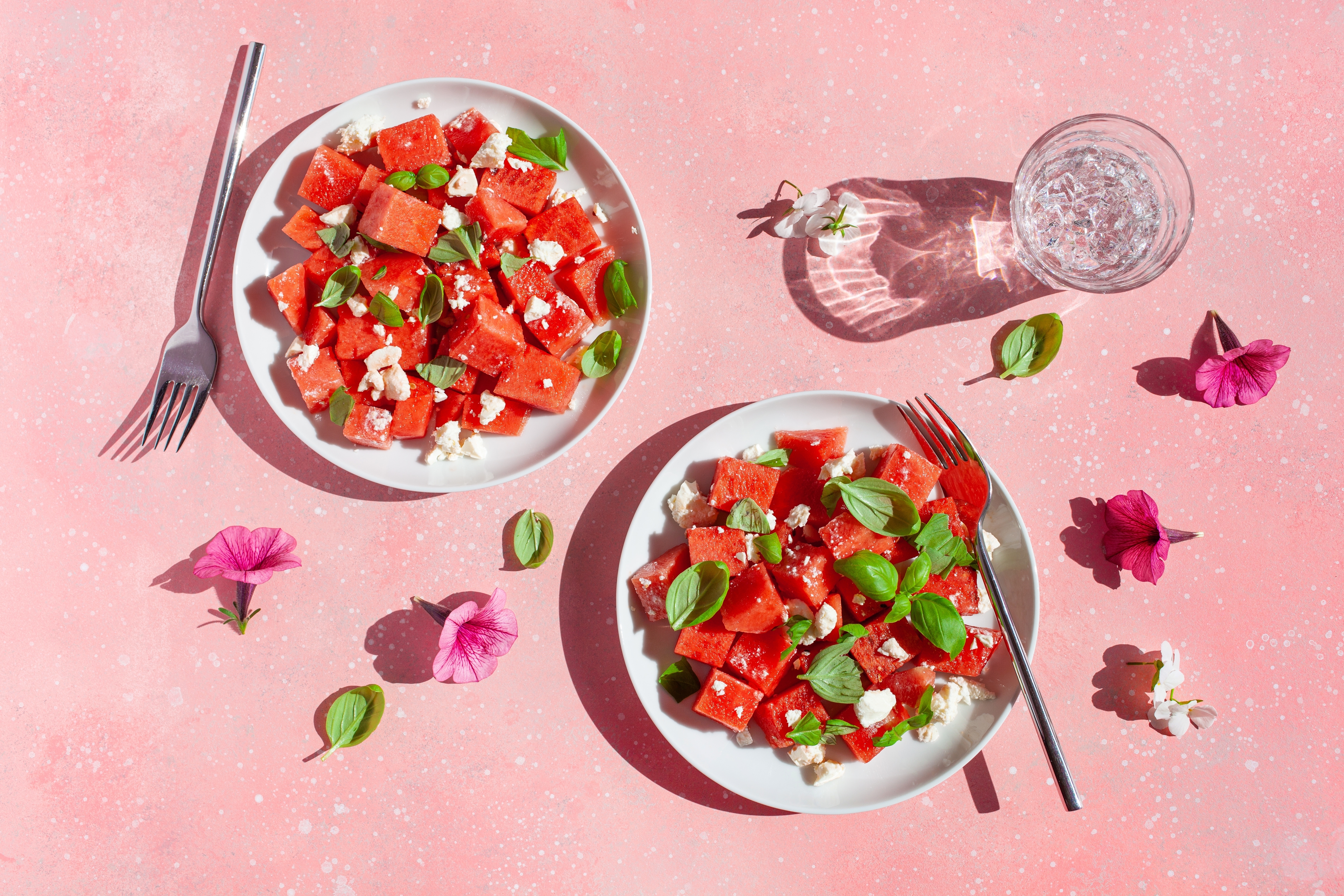 Two white plates with chopped watermelon, crumbled white cheese, and fresh basil leaves are arranged on a pink splattered background. A fork on each plate, a glass of water, and scattered flowers and basil leaves decorate the scene.